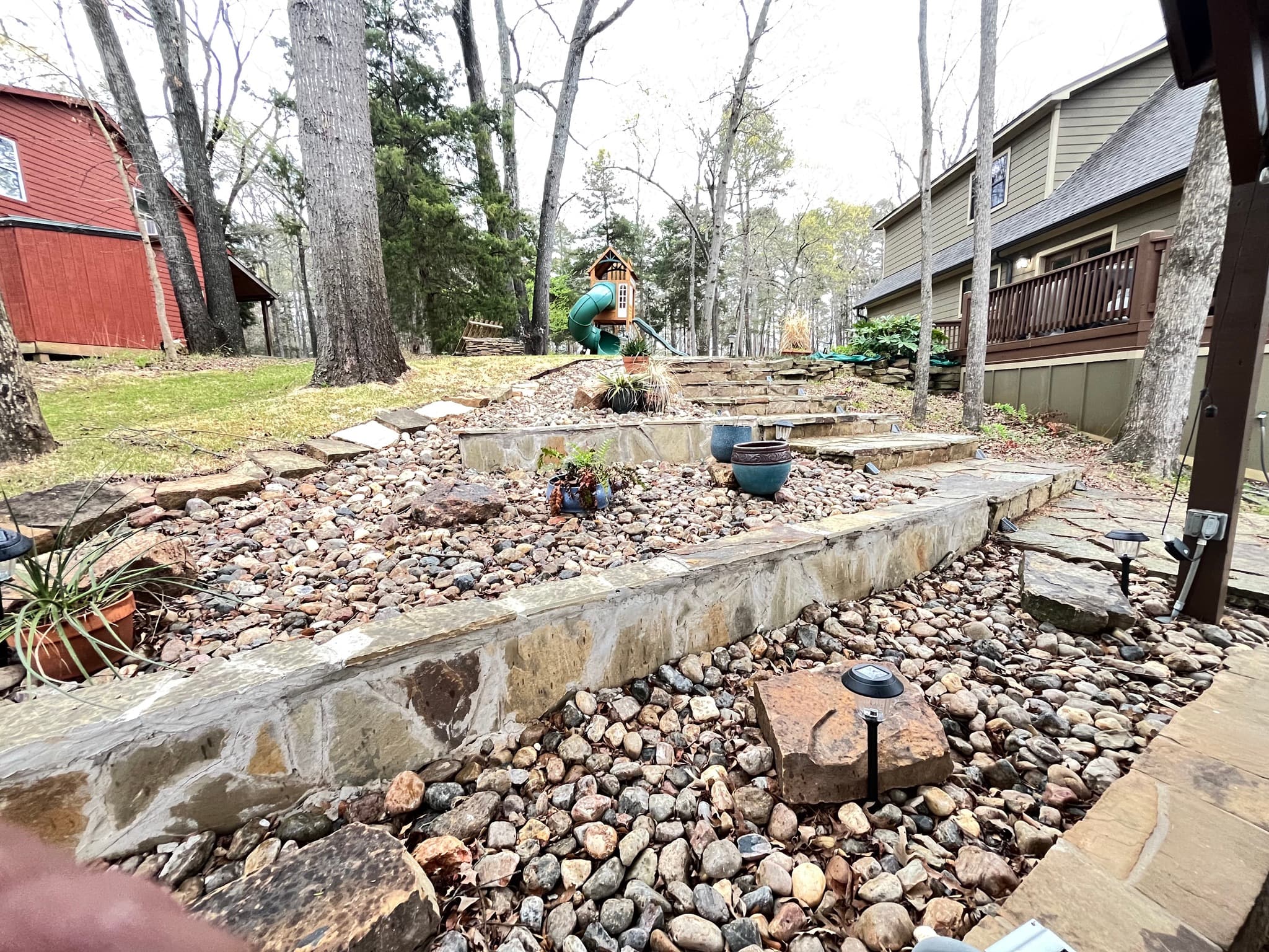 Tiered stone landscape with river rocks and potted plants leading to a backyard playset.