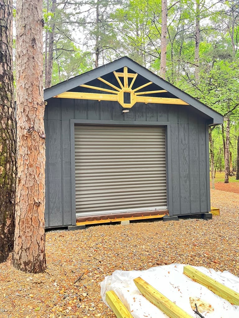 Dark grey shed with roll-up door and wooden sunburst gable decoration in the woods.
