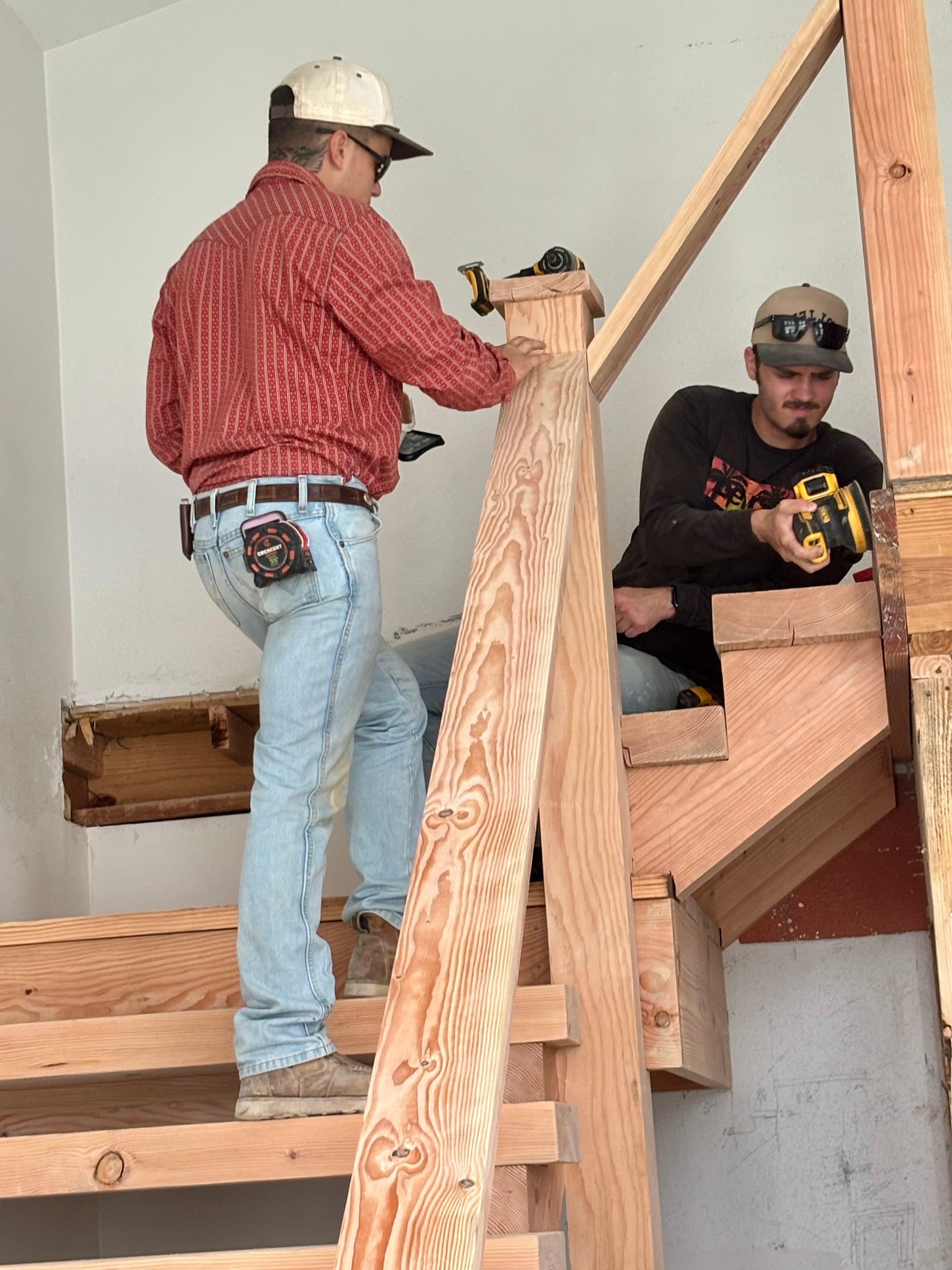 Two construction workers in hats use power tools to build a wooden staircase indoors.